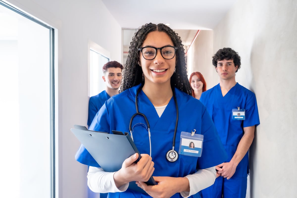 Group of diverse medical students or young healthcare professionals walking in a hospital corridor, focused female nurse or doctor smiling at camera with clipboard and stethoscope