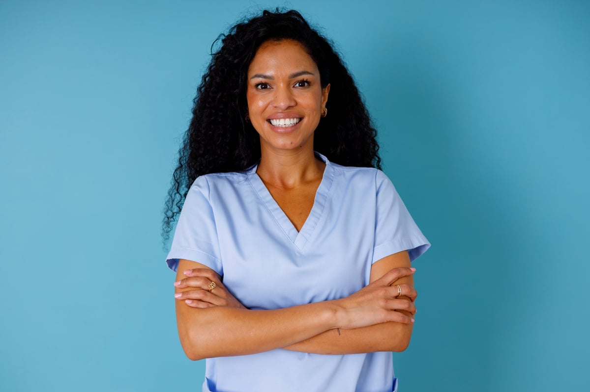 Young adult woman medical professional wearing scrubs, smiling directly at the camera with her arms crossed, portraying confidence and care