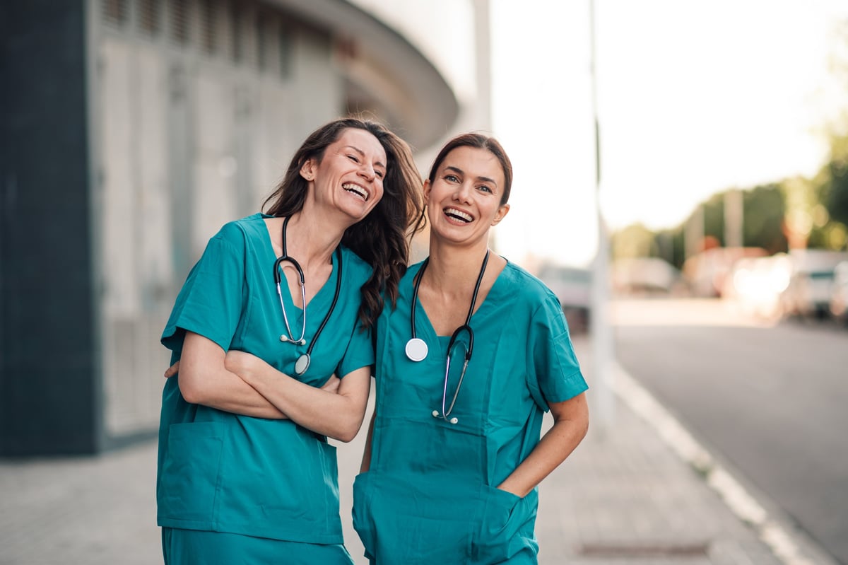 Two cheerful young adult healthcare professionals smiling and laughing, standing outdoors in their teal scrubs and stethoscopes