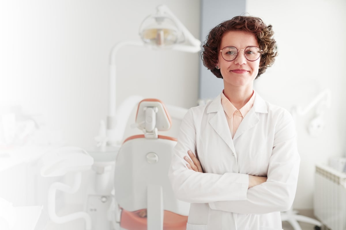 Successful smiling middle-aged dentist in eyeglasses with crossed arms standing in own office