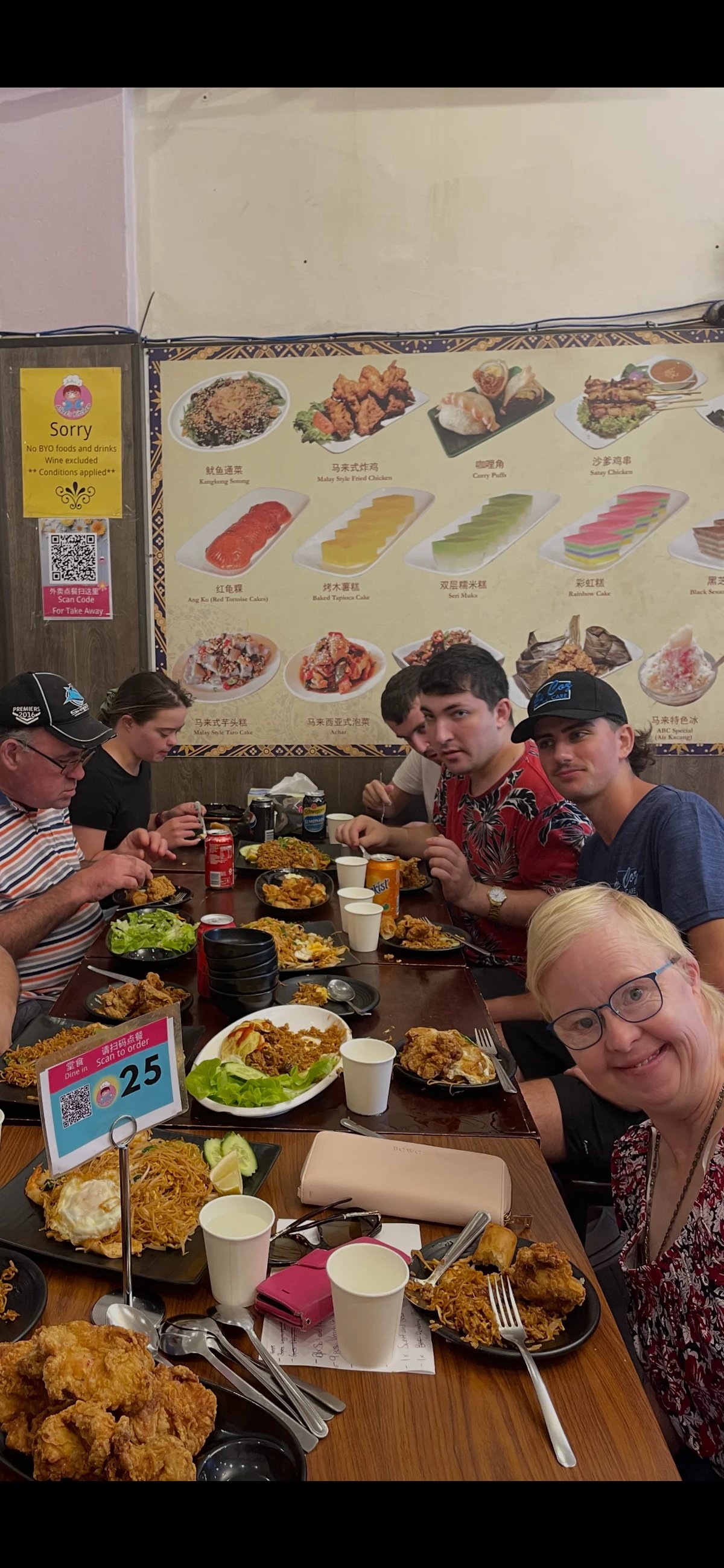 Group of five people dining together at an Asian restaurant with various dishes and drinks on the table, menu visible on wall behind them