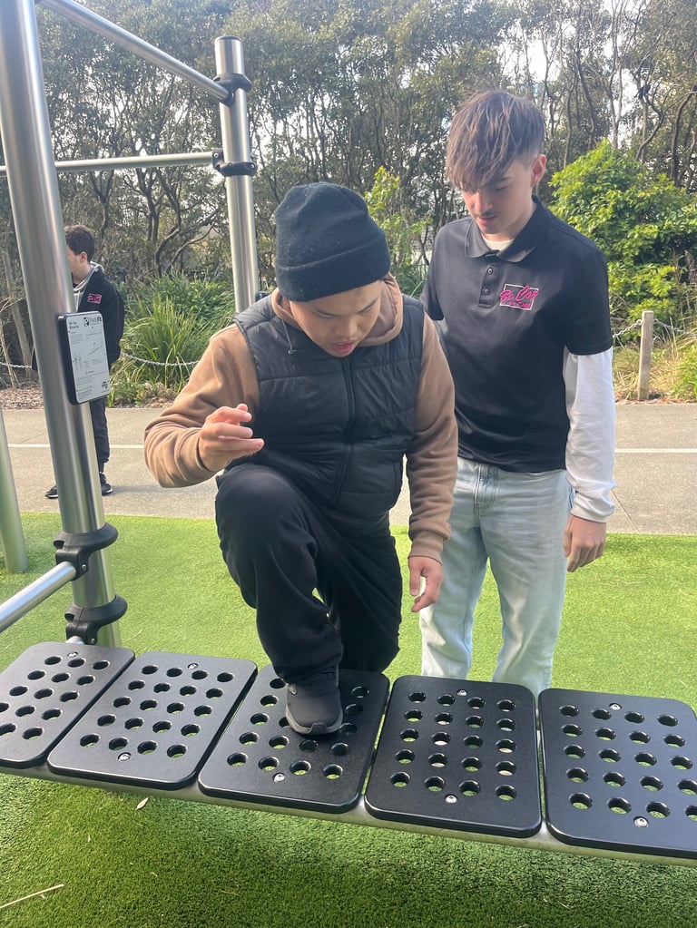 Person bending down to use outdoor exercise equipment with instructor standing nearby and trees in background