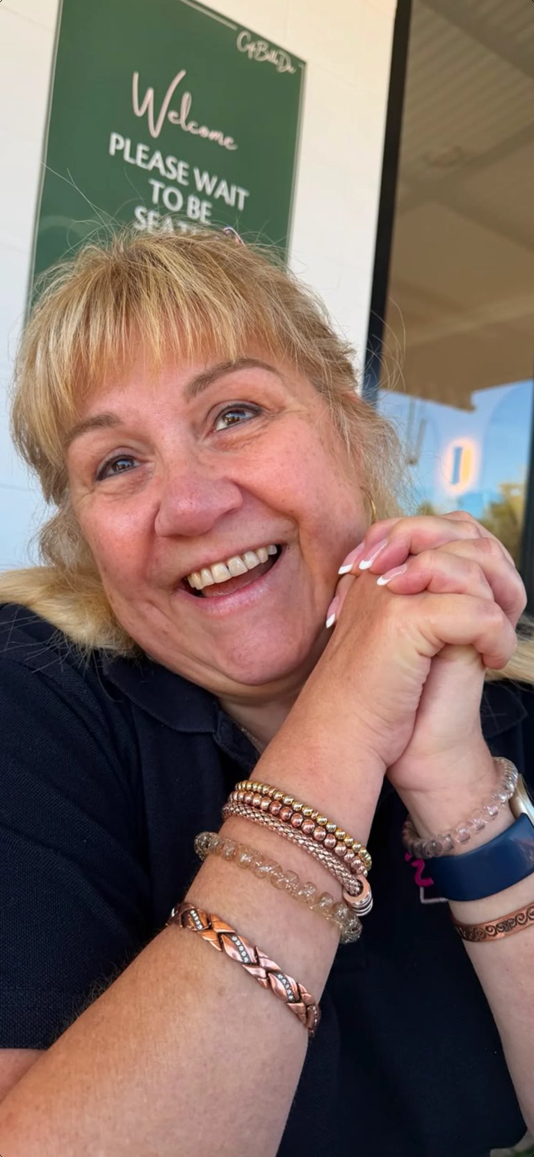 Smiling woman with short blonde hair and multiple bracelets posing indoors near a green welcome sign