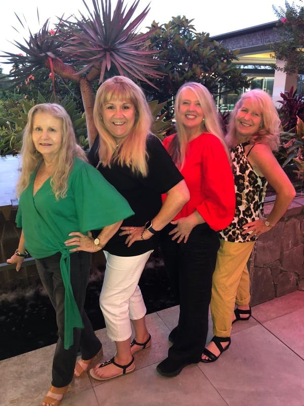 Four women posing together outdoors at a tropical garden venue with colorful plants and buildings in background