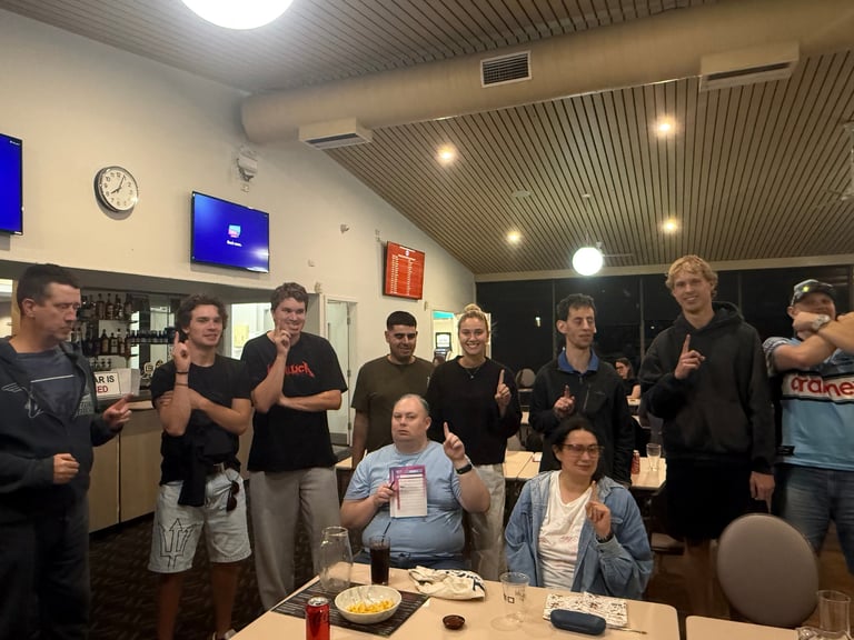 Group of people gathered in an indoor space, mostly wearing black clothing, standing around a table with food and drinks