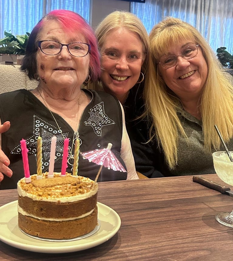 Three women smiling at a birthday celebration with a cake displaying lit candles in front of them