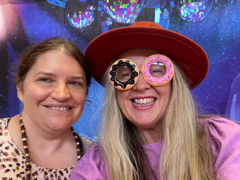 Two women smiling at a party with colorful disco ball backdrop. One wears a red hat and novelty donut and star sunglasses