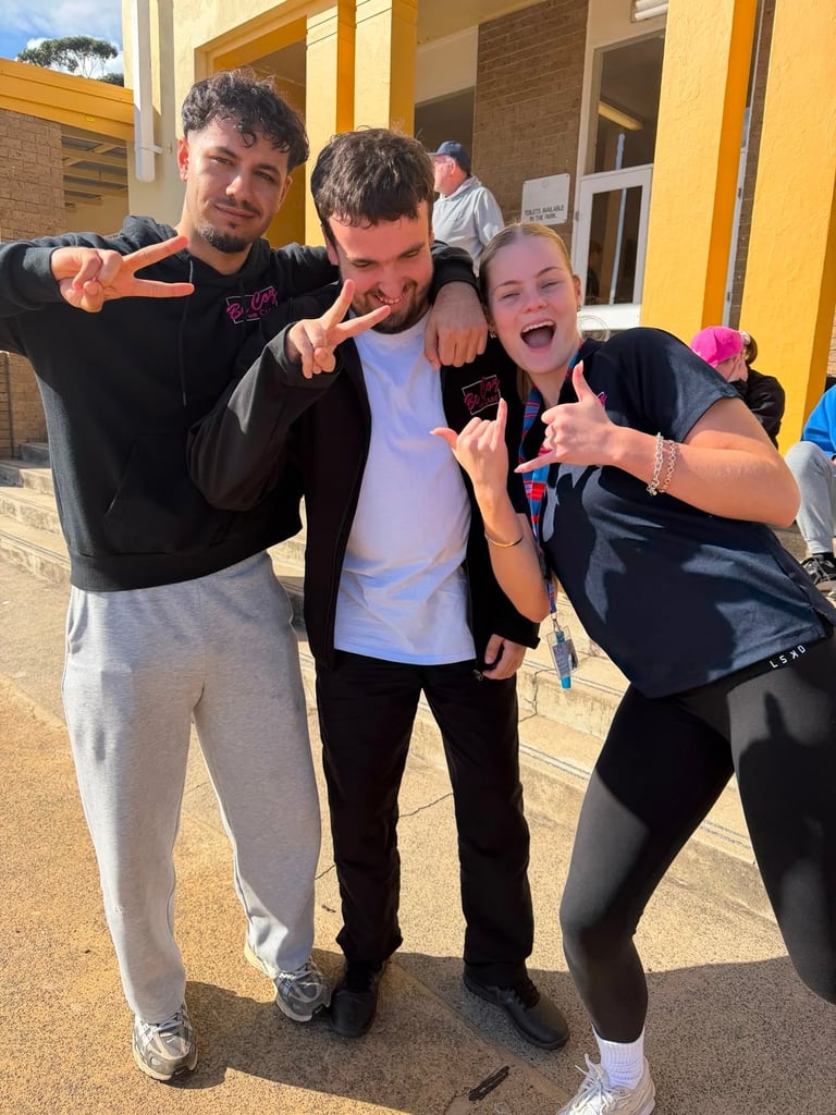 Three young people posing outdoors with arms around each other, making playful hand gestures and smiling at the camera in front of a yellow building.