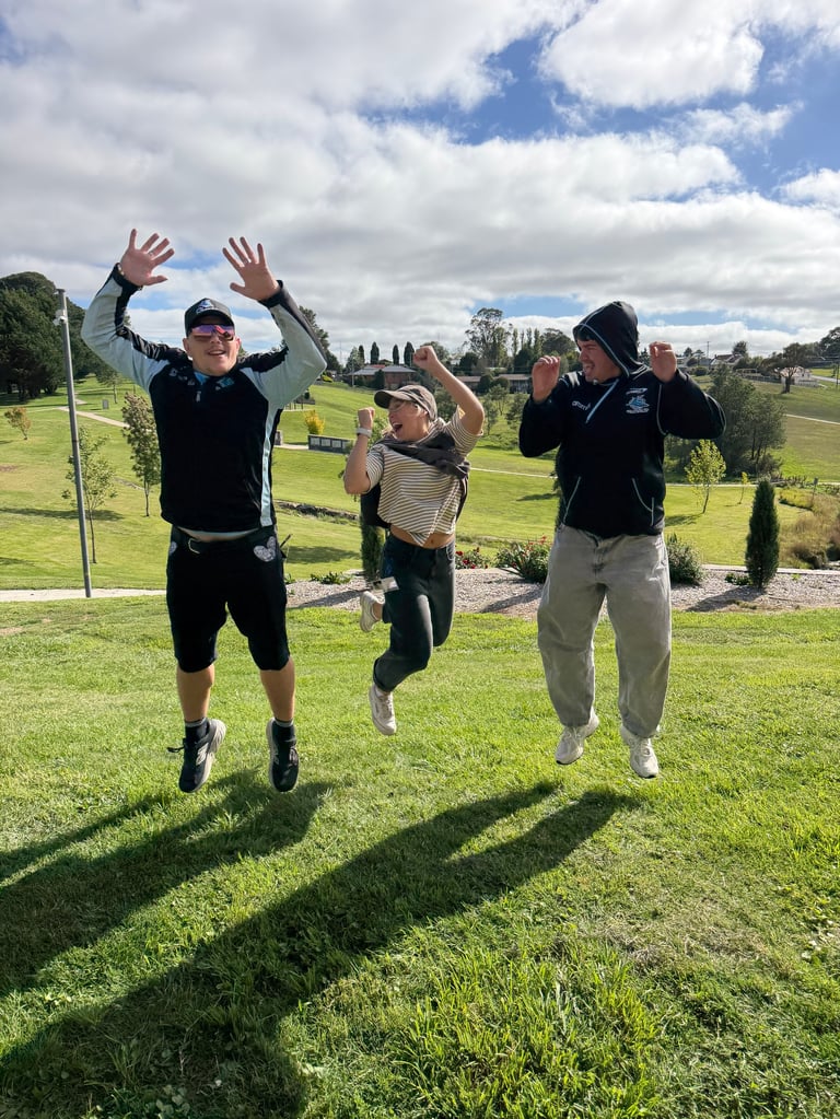 Three people jumping with hands raised on a green field under a blue sky with white clouds