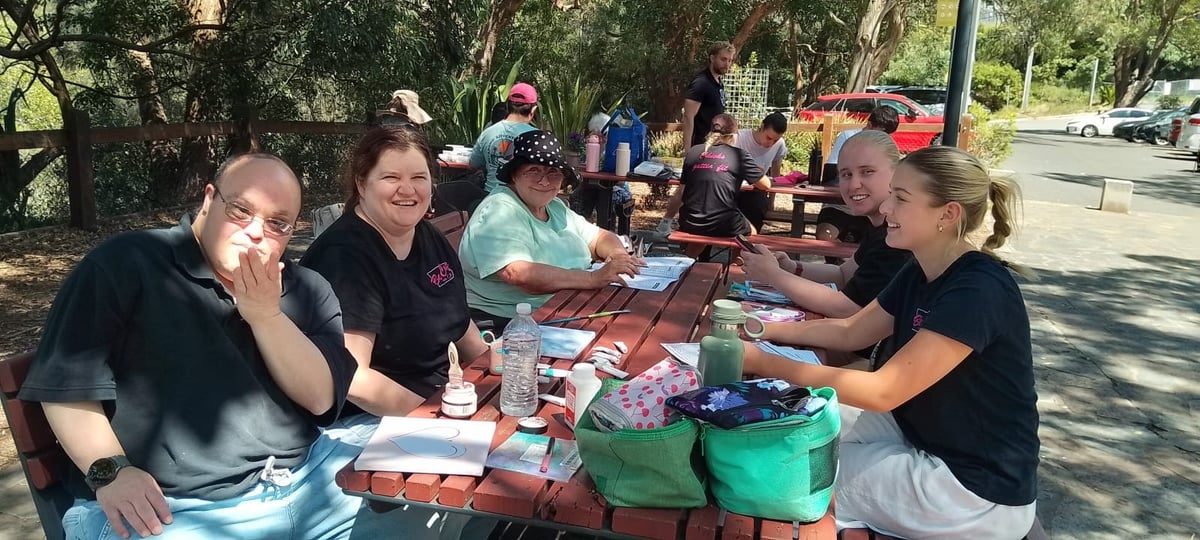 Group of people seated at a picnic table outdoors during daytime, with food and drinks, trees and parked cars visible in background