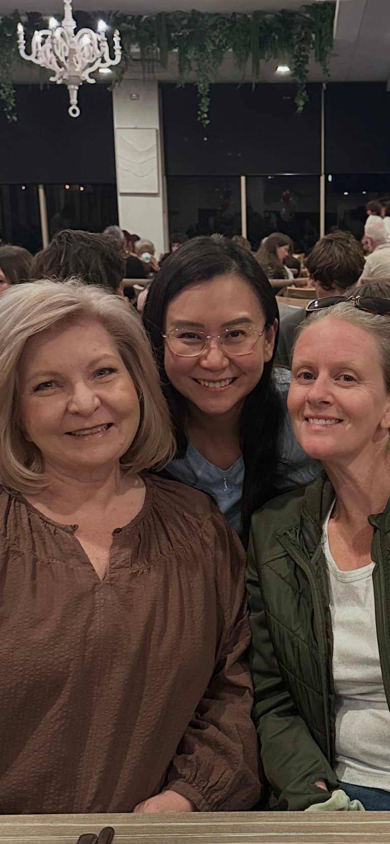 Three smiling women at an indoor event with a white chandelier and hanging plants visible in the background