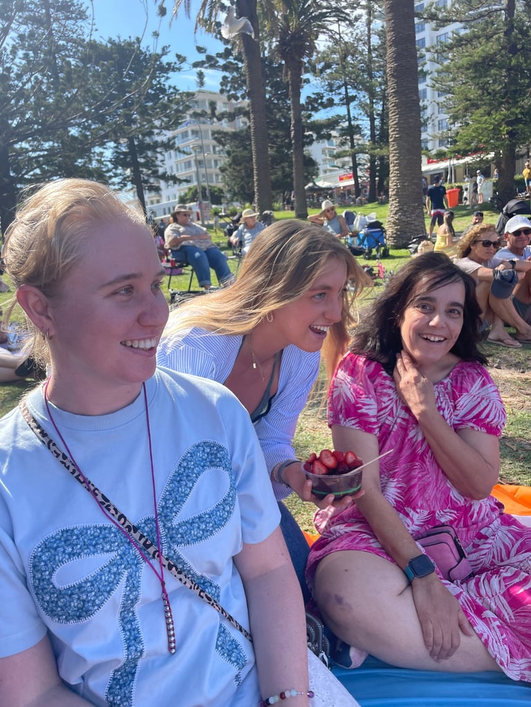 Three smiling women sitting together at an outdoor park picnic with tall trees, grass, and buildings visible in the background