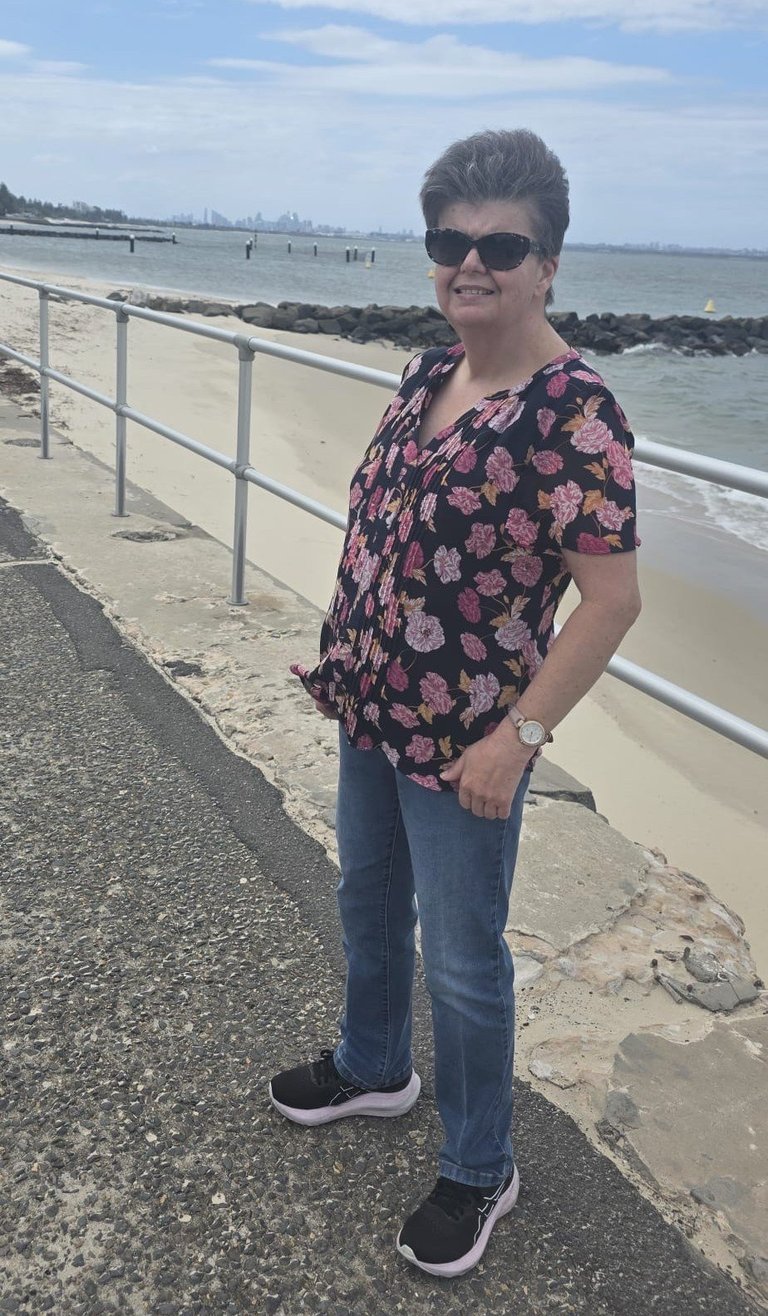 Woman with short dark hair wearing sunglasses, a black floral top, and jeans standing on a beach promenade with ocean and rocky breakwater in the background