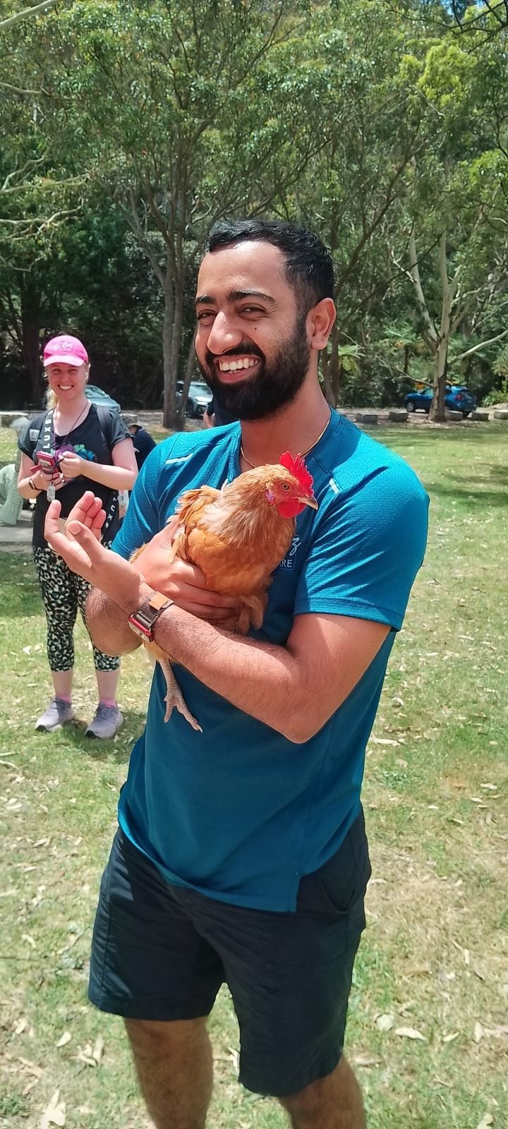 Man in blue shirt holding a brown hen, smiling at an outdoor park gathering with trees and other people in the background