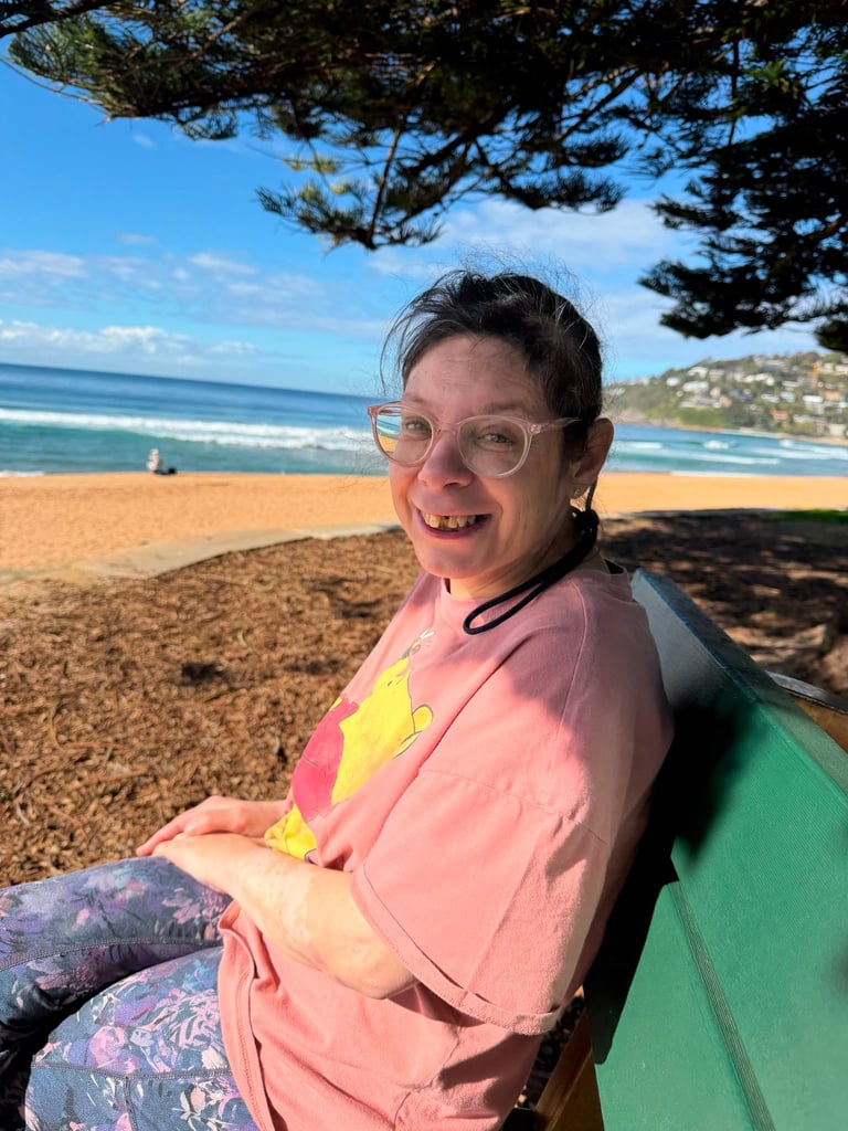 Smiling person wearing glasses and pink shirt sitting on beach bench with ocean and trees in background