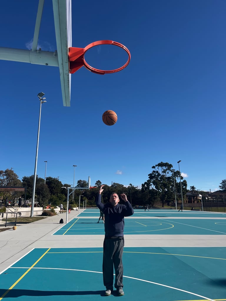 Person shooting basketball on outdoor court under clear blue sky, ball in mid-air toward hoop