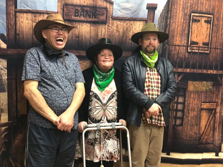 Three people in cowboy hats and western attire posing in front of a wooden bank building set, one person holding a wheelbarrow