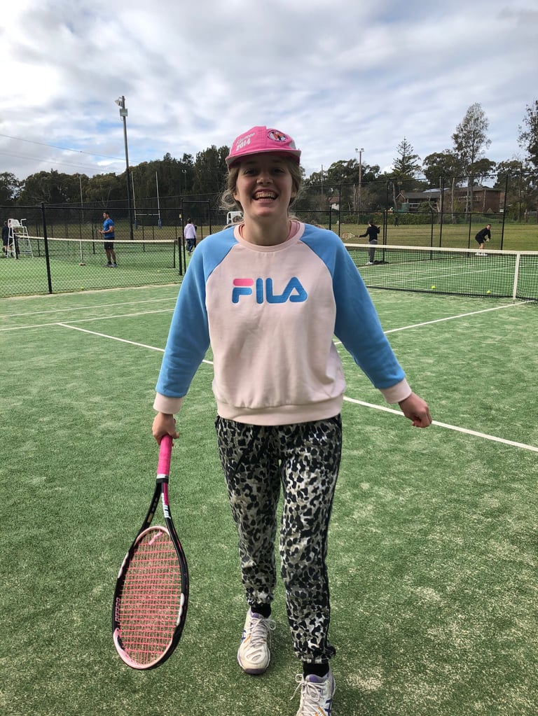 A smiling person wearing a pink helmet and FILA sweatshirt holds a tennis racket on an outdoor green court with other players visible in the background