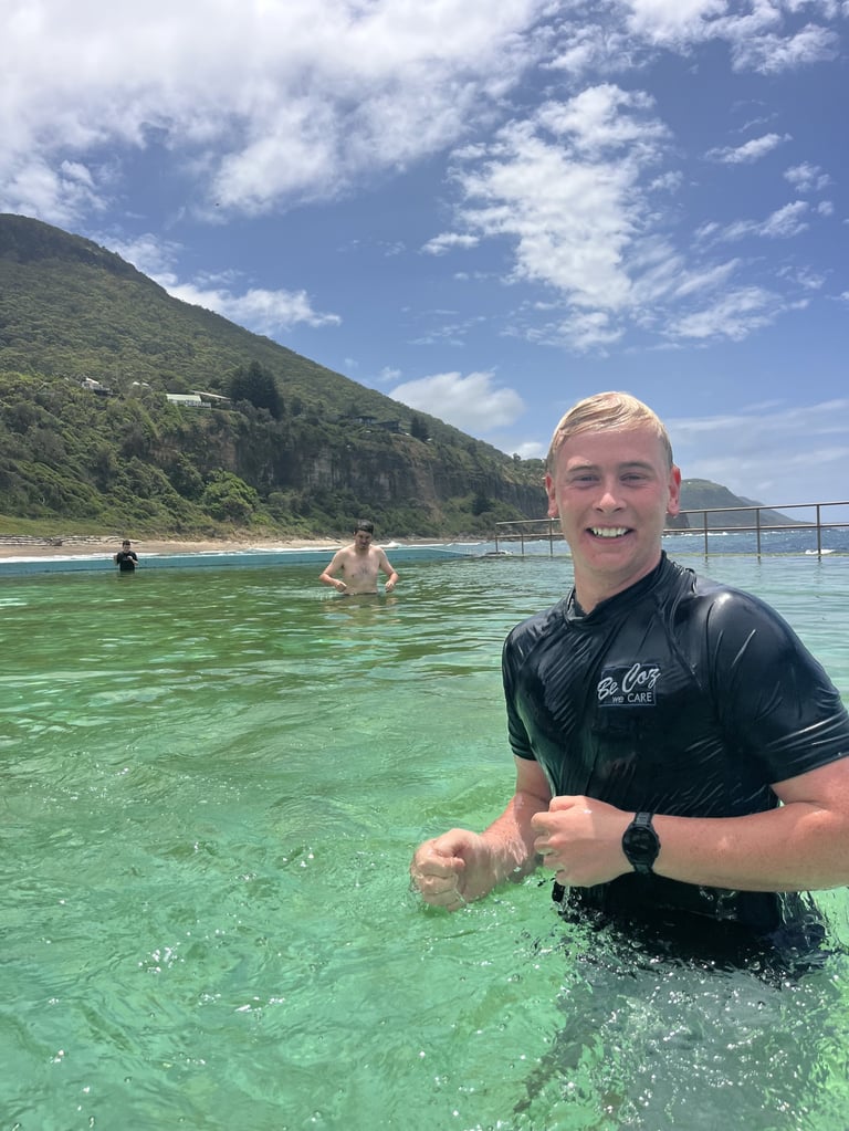 Young man in black rash guard smiling in turquoise ocean water with green mountains and beach in background