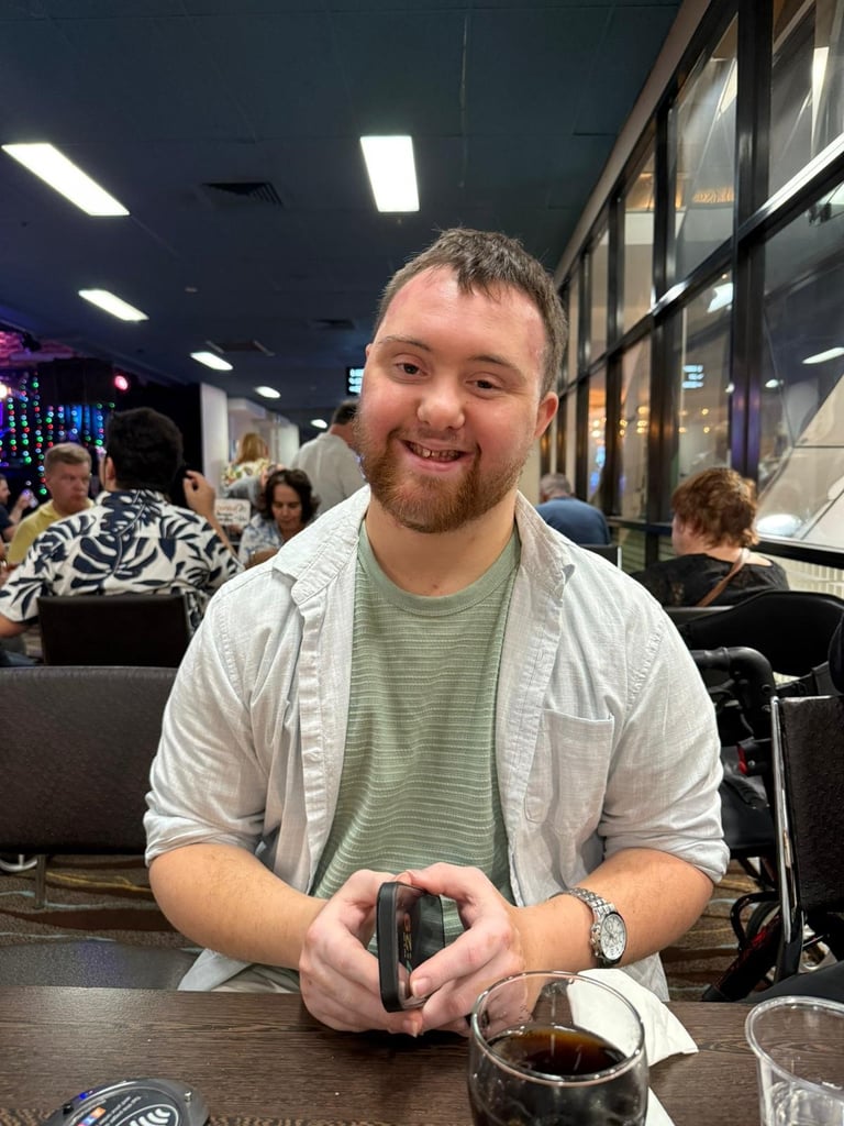 A smiling man in a white shirt and green striped shirt holds a video game controller at an indoor venue with other people in the background