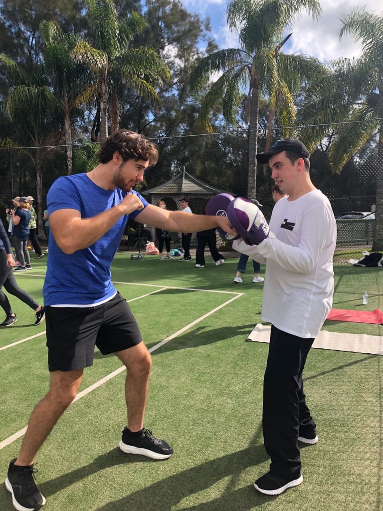 Two men fist bumping during outdoor boxing training session in a park with palm trees and spectators in background