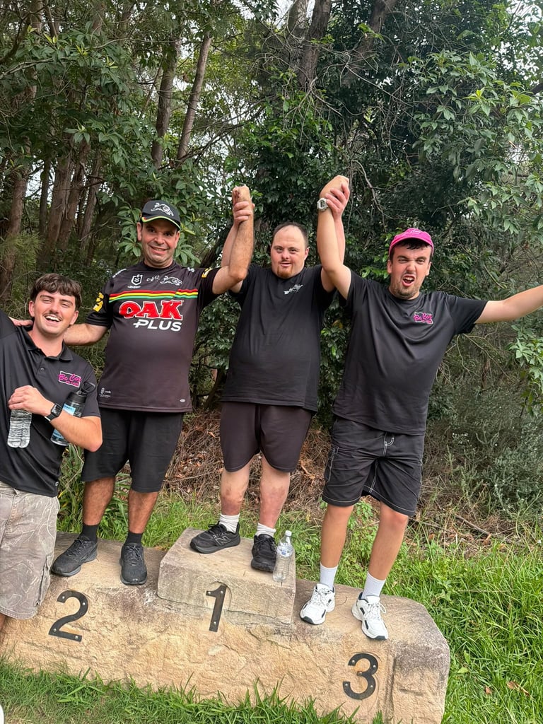 Four men posing on a podium with numbers 2, 1, and 3, wearing dark shirts, celebrating outdoors with trees in background
