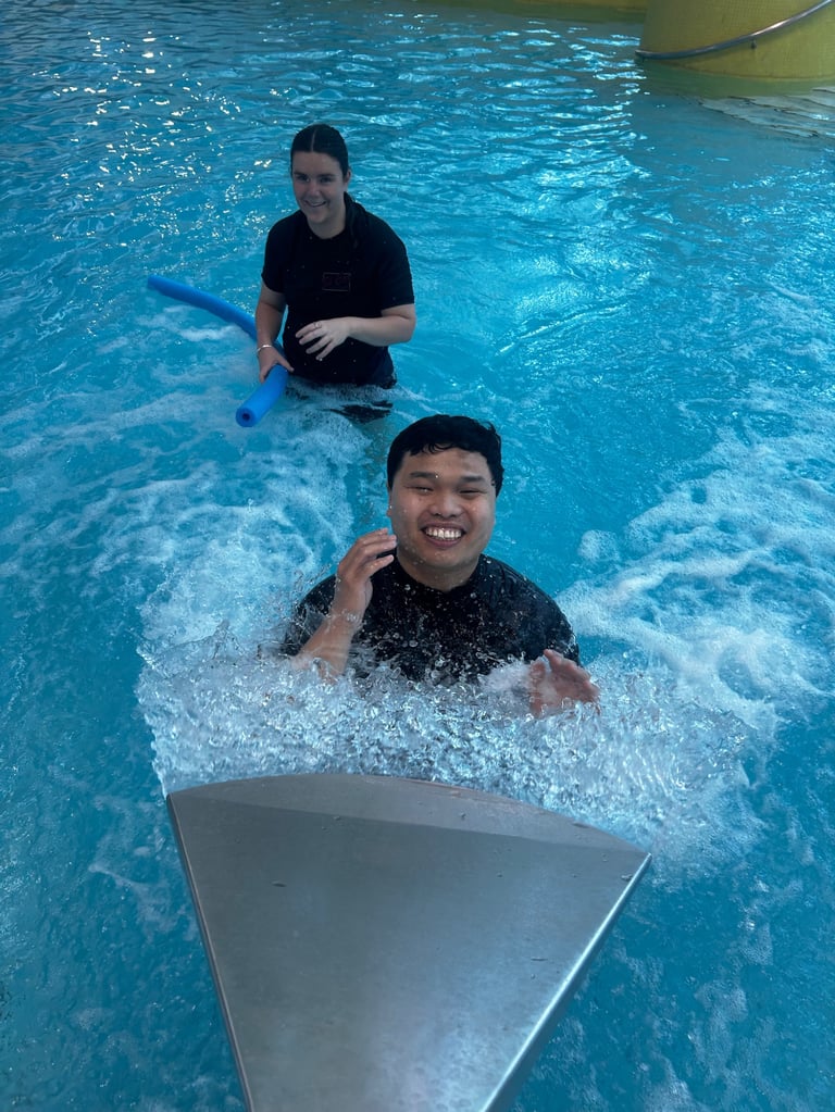 Two people in a swimming pool during a swimming lesson, with an instructor assisting a smiling student on a kickboard