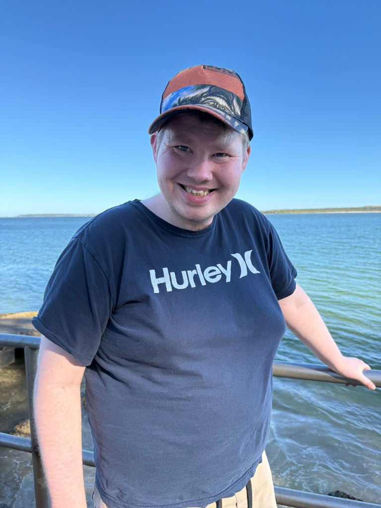 Smiling young person wearing a Hurley shirt and red cap standing at a dock with blue ocean and clear sky in background