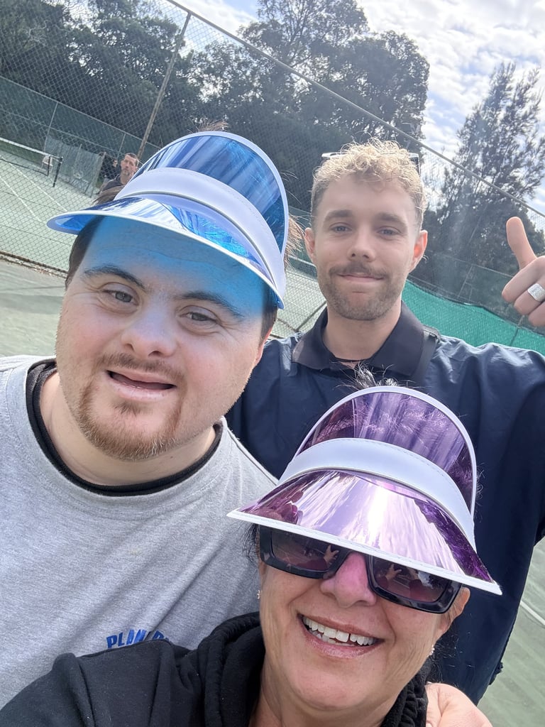 Three people wearing colorful visors smile at a tennis court with trees in the background