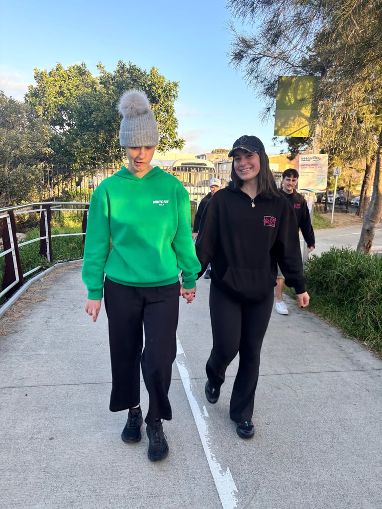 Two young women walking together on a sidewalk, one wearing a green hoodie and gray beanie, the other in a black hoodie