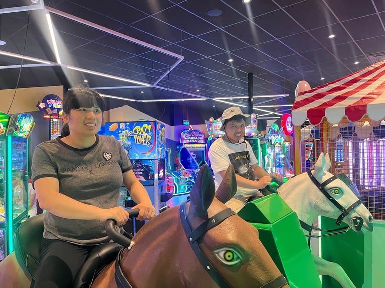 Two people riding mechanical arcade games in a colorful arcade facility with neon lights and game machines