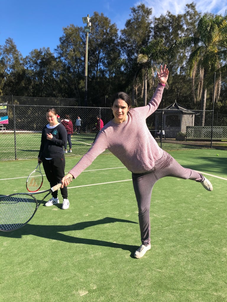 Woman in pink sweater posing playfully on tennis court holding a racket with instructor in background