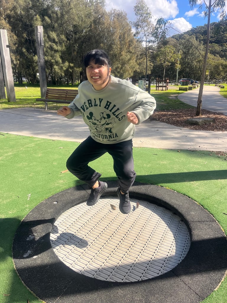 Person jumping on a trampoline in a park wearing a Beverly Hills California sweatshirt and black pants, with green lawn and trees in background