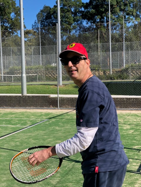 Man in red cap and sunglasses holding tennis racket on outdoor court with metal fence and trees in background
