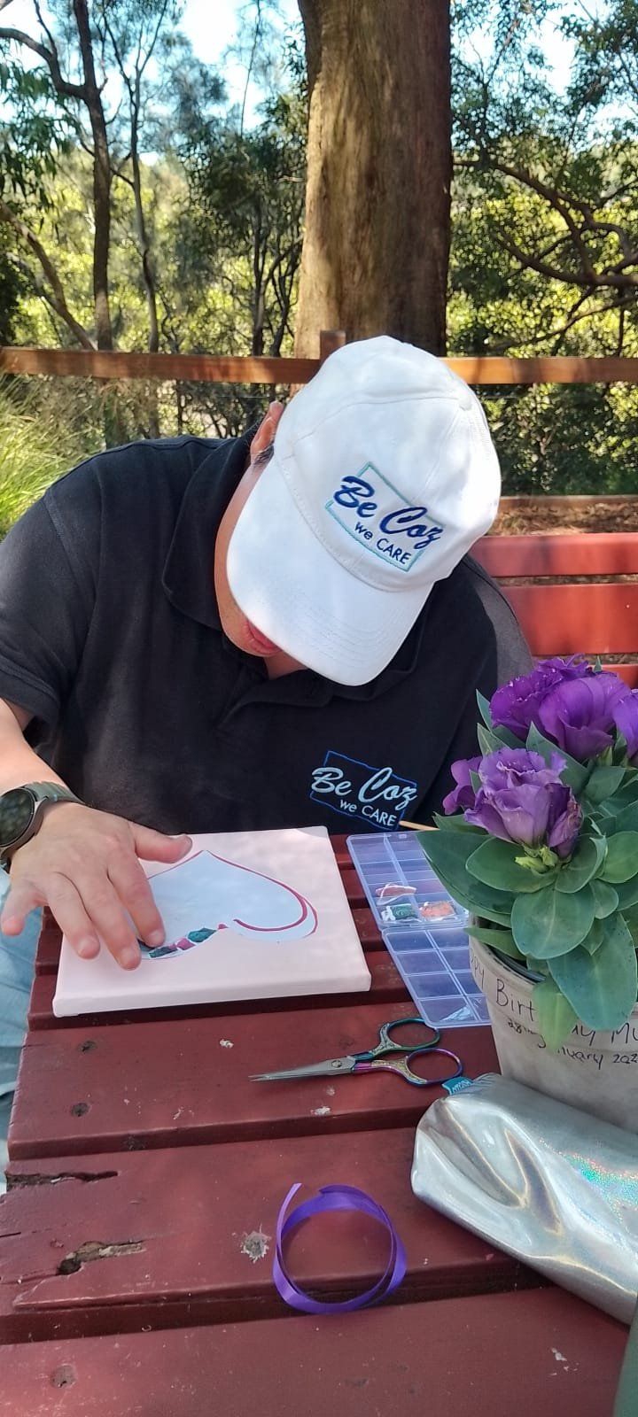Person in Be Coz Care branded cap and shirt working on an art project at an outdoor wooden table with purple flowers and craft supplies