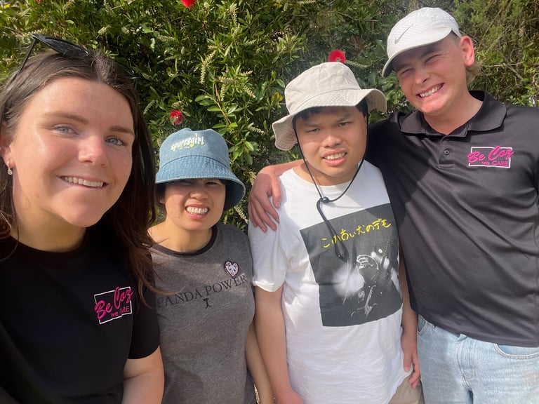 Four smiling teenagers standing together outdoors in front of green foliage wearing casual clothing and white and blue caps