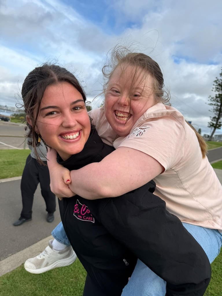 Two young women smiling and hugging outdoors on a grassy area with a road and cloudy sky in the background