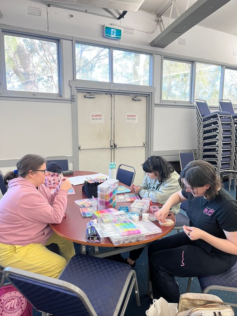 Four students working together at a round table with colorful craft materials in a bright classroom with large windows and stacked chairs