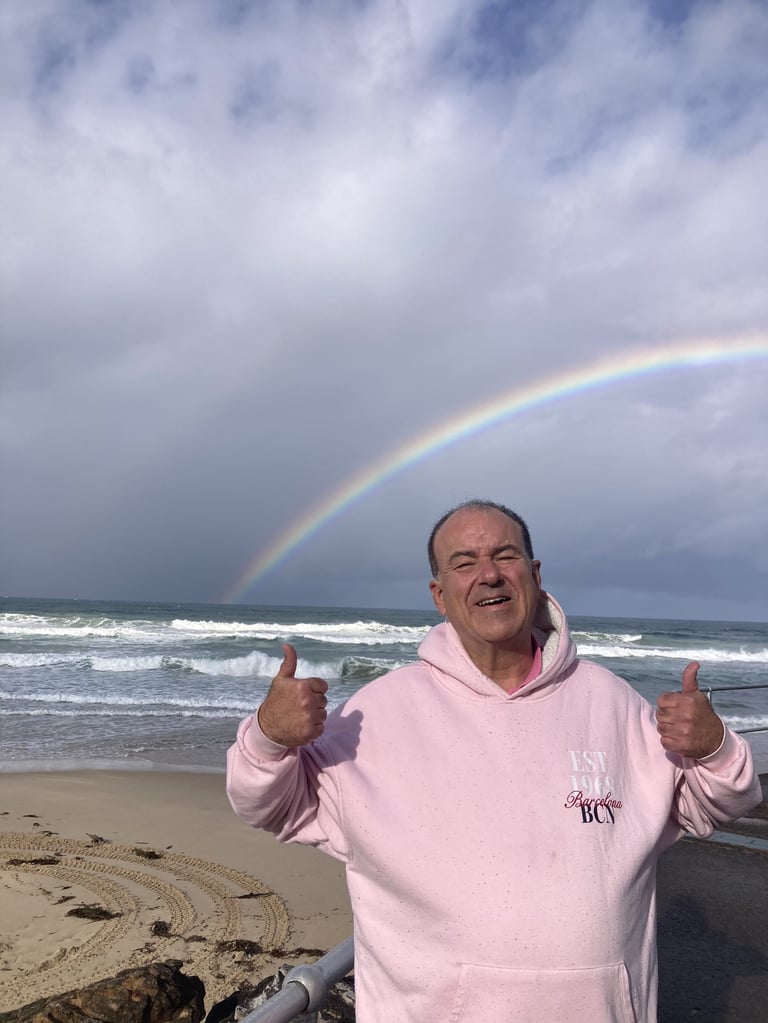 Man in pink hoodie giving thumbs up on beach with rainbow and ocean waves in background