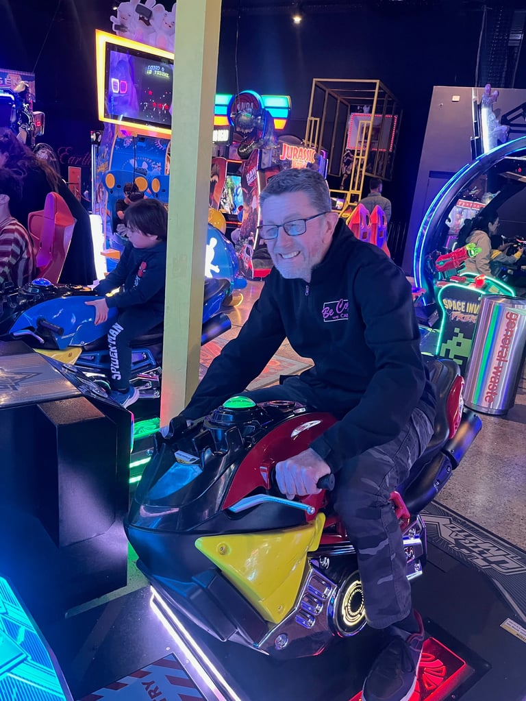 Man smiling on colorful racing arcade game in neon-lit arcade venue surrounded by other games and players