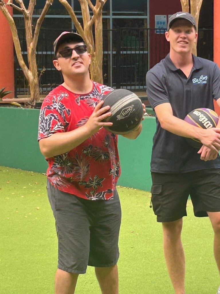 Two men holding basketballs on a bright green lawn, with bare tree branches and a colorful building visible behind them