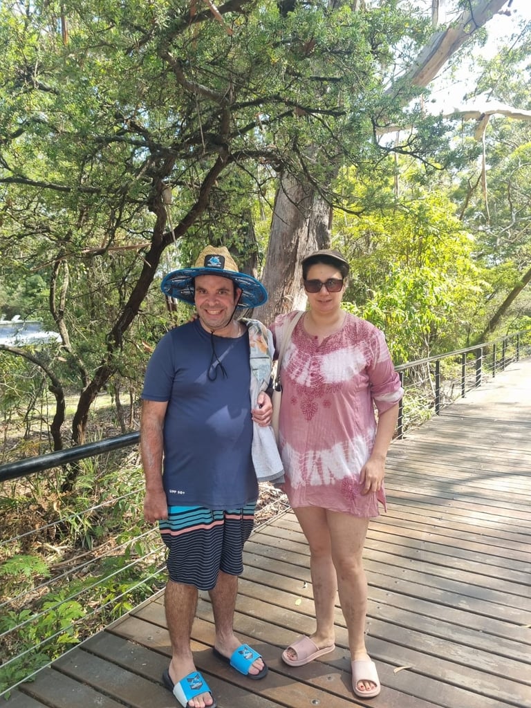 Two people posing on a wooden bridge surrounded by large green trees in a forest setting