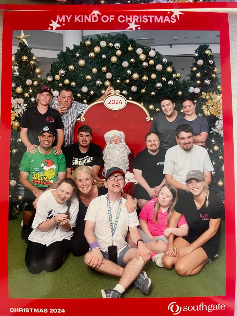 Group photo with Santa at Christmas 2024 celebration, large decorated Christmas trees with gold ornaments in background, red framed photo setup with My Kind of Christmas text