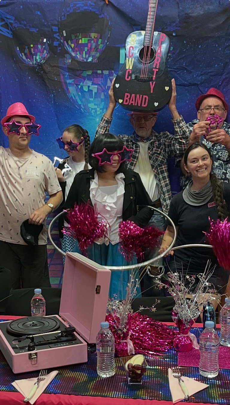 Group of five people posing at a music-themed party with props including star-shaped glasses, a colorful guitar, pink fedora hats, and a pink turntable on the table.