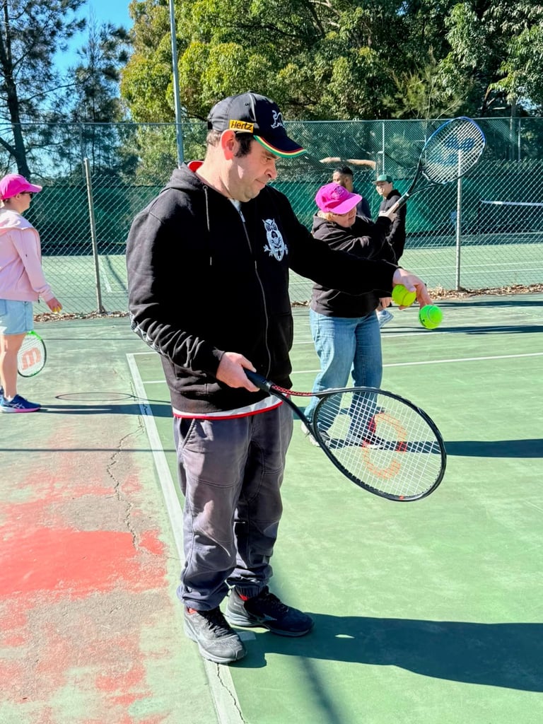 An instructor in black jacket and cap demonstrates tennis technique holding a racket on a colorful outdoor court with children watching
