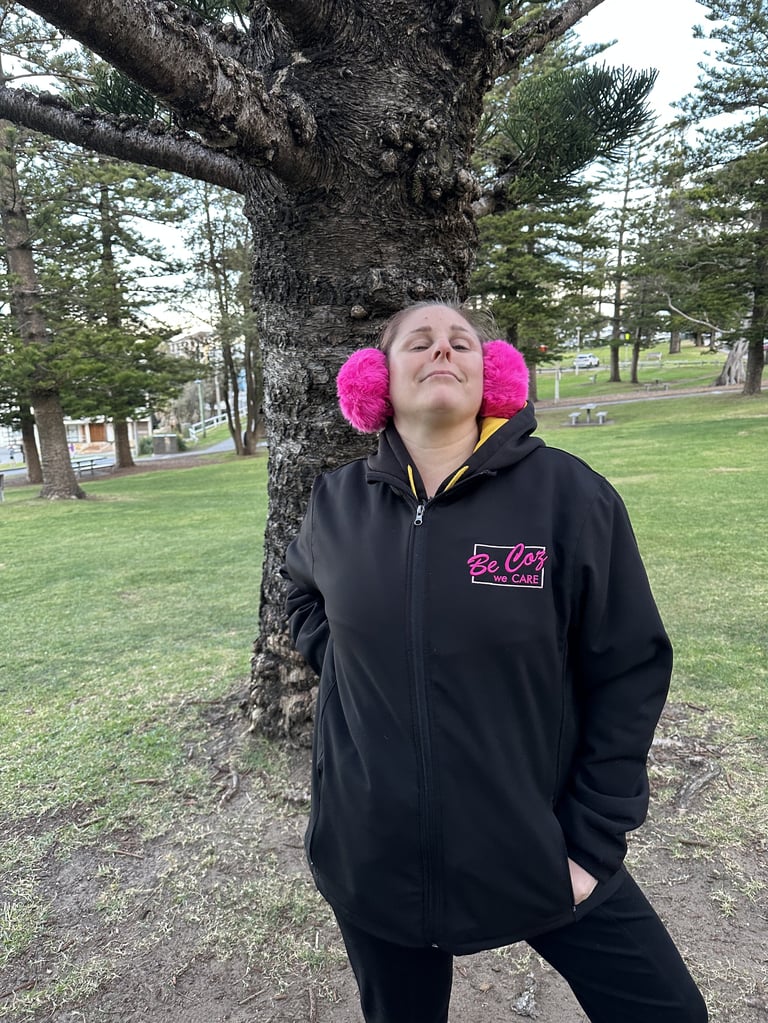 Person wearing black hoodie with pink fuzzy earmuffs leaning against large tree in cemetery park
