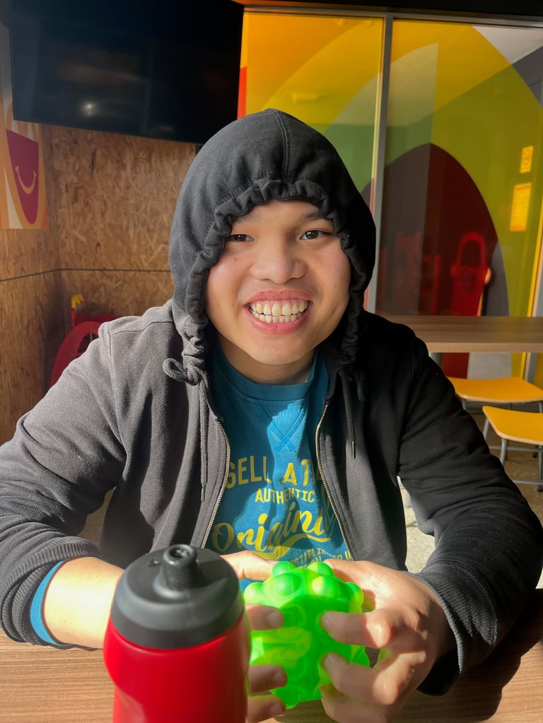 Young person wearing a black hoodie and gray jacket, smiling while holding a green toy, with a red bottle nearby