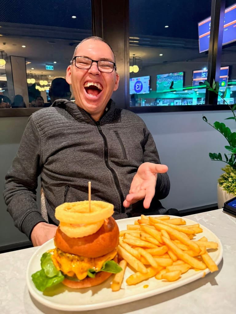 Man wearing glasses and gray hoodie laughing while pointing at a large burger with fries at a restaurant table