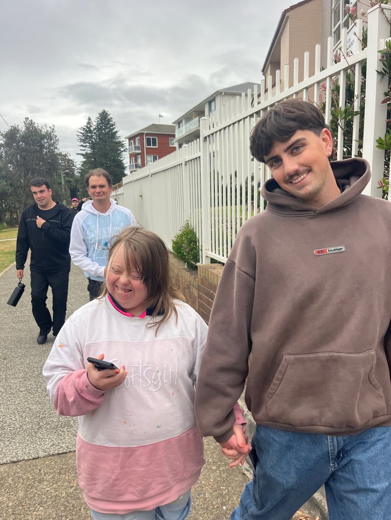 A smiling teen boy in a brown hoodie holds hands with a younger girl in pink while two adults walk behind them on a residential driveway